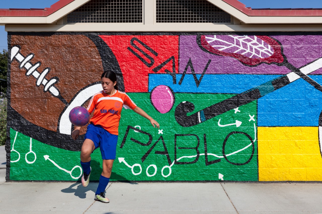 一名青少年足球运动员在拉姆里尔体育公园的壁画前玩着球。在圣巴勃罗工业区新建的拉姆里尔体育公园(Rumrill Sports Park)，足球队将在三块有人造草皮和球场灯光的新场地上进行比赛。圣巴勃罗的儿童肥胖率是全县最高的，但公园和开放空间却最少。该市和圣巴勃罗经济发展公司(San Pablo Economic Development Corp.)与联邦和州机构合作，修复空置的铁路用地，然后通过联邦新市场税收抵免计划筹集资金，建造最先进的油田。“我们在这里没有一个安全的、有照明的、有衬里的、有草皮的地方。最近的一个在伯克利，”圣巴勃罗经济发展公司总经理Leslay Choy说。