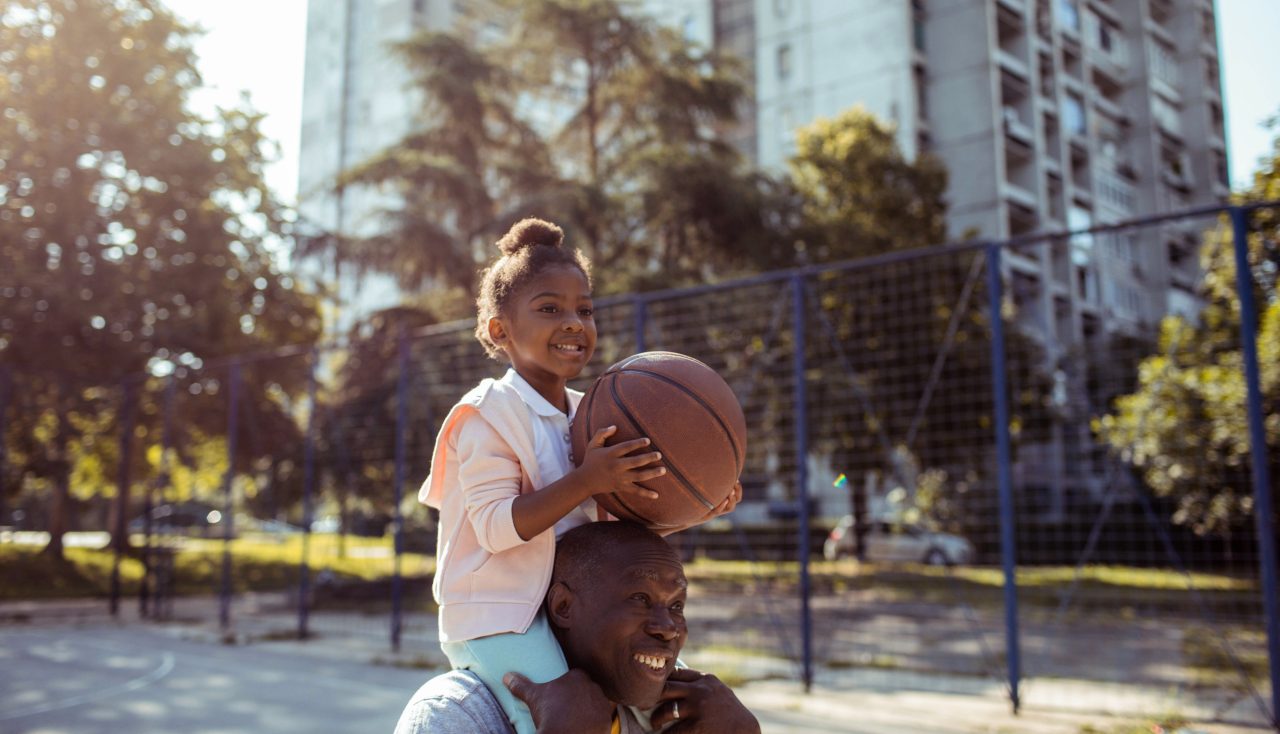 一个年轻女孩坐在一个男人的肩膀上，手里拿着一个篮球。